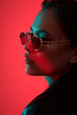 Kıyafetler, aksesuarlar, gözlükler. Çekici, kendine güvenen esmer kız şık güneş gözlükleriyle poz veriyor ve kırmızı stüdyo ışıklarında siyah deri ceket giyiyor. Kadın modası.