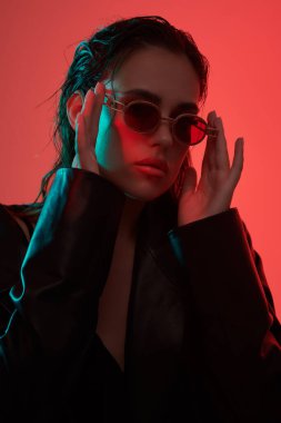 Çekici, kendine güvenen esmer kız şık güneş gözlükleriyle poz veriyor ve kırmızı stüdyo ışıklarında siyah deri ceket giyiyor. Kadın modası. Giysiler, aksesuarlar, gözlükler..