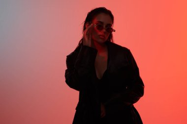 Çekici, kendine güvenen esmer kız şık güneş gözlükleriyle poz veriyor ve kırmızı stüdyo ışıklarında siyah deri ceket giyiyor. Kadın modası. Kıyafetler, aksesuarlar, gözlükler. Metin için yer.