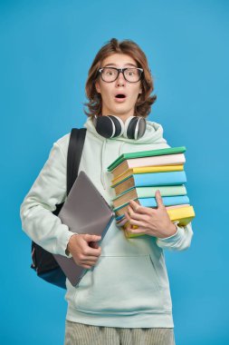 Modern bir lise öğrencisi ders kitapları ve bir dizüstü bilgisayar bulundurur ve çok şaşırır. Modern eğitim. Mavi stüdyo arka planı.