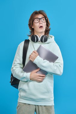 Duygusal genç liseli çocuk elinde bir dizüstü bilgisayar tutuyor ve şaşkınlıkla ağzını açıyor. Eğitici bir kavram. Mavi stüdyo arka planı.