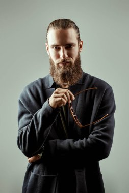 Gri bir stüdyo arka planında şık bir hırka ve şık güneş gözlüğü takmış sakallı ve bıyıklı yakışıklı bir adamın portresi. Erkekler için tarz ve moda.