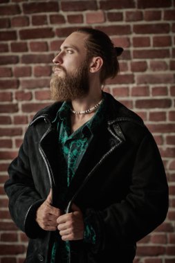 Sakallı, bıyıklı, şık bir gömlek ve deri ceketle tuğla duvar arkasında poz veren yakışıklı, vahşi bir adamın portresi. Erkek modası ve güzelliği.