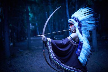 Ulusal kostüm ve başlık giymiş Amerikalı bir Hintli kız elinde bir yayla ormanda duruyor. Etnik Amerikan kültürü. Ay ışığı..