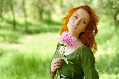 Fantezi kadın kahraman. Çekici kızıl saçlı bir kız yaz yeşilliğinin arka planında büyük bir şakayık ile duruyor. Kelt kültürü. Kadın güzelliği ve çiçekleri. Boşluğu kopyala.