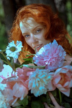 Çekici kızıl saçlı bir kız elinde büyük bir buket şakayıkla yaz yeşilliğinin arka planında duruyor. Kadın güzelliği ve çiçekleri. 