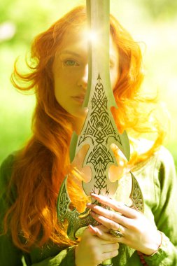 Fantezi kadın kahraman. Muhteşem uzun kızıl saçlı, tarihi bir Kelt elbisesi olan genç ve güzel bir savaşçı kadın bir ormanda elinde kılıçla duruyor. Orta Çağ Biçimi. 