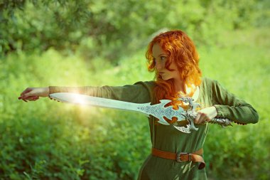 Muhteşem uzun kızıl saçlı, tarihi bir Kelt elbisesi olan genç ve güzel bir savaşçı kadın bir ormanda elinde kılıçla duruyor. Orta Çağ. Fantezi. Tarihi yeniden yapılandırma.