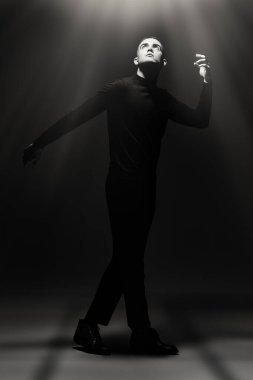 Siyah elbiseli genç bir adamın sanat portresi. Karanlıkta duran ve yüzünü aydınlatan bir ışık huzmesi. Siyah arkaplana karşı tam boy siyah beyaz portre. Psikoloji. Tiyatro.