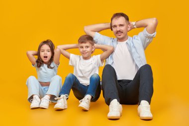 Mutlu bir baba, sevgili oğlu ve kızıyla yerde oturup kas egzersizleri yapıyor. Mutlu aile ve spor. Sarı stüdyo arka planında tam boy portre.