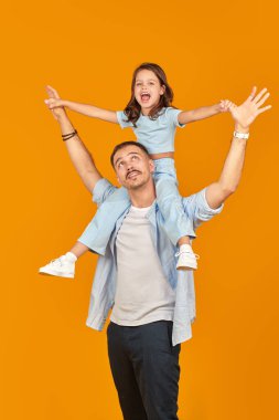 Mutlu babasının boynunda oturan neşeli küçük bir kızın aile portresi. Sarı arka planda stüdyo çekimi.