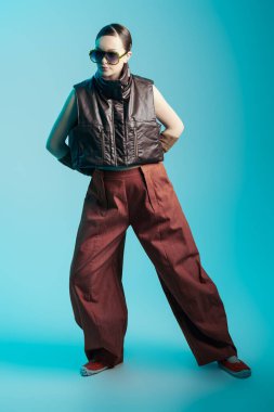Mavi stüdyo arka planında bahar-yaz koleksiyonundan şık tasarımcı kıyafetleri giymiş bir manken kızın tam boy portresi. Yüksek moda çekimi. Moda güneş gözlüğü.