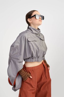 Gri ceketli, geniş kahverengi pantolonlu ve gri arka planda aynalı güneş gözlüklü güzel bir mankenin portresi. Fütürist moda tarzı. Yüksek moda.