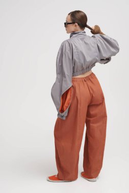 Gri ceketli, geniş kahverengi pantolonlu ve gri arka planda aynalı güneş gözlüklü güzel bir kız modelin tam boy portresi. Fütürist moda tarzı. Yüksek moda.