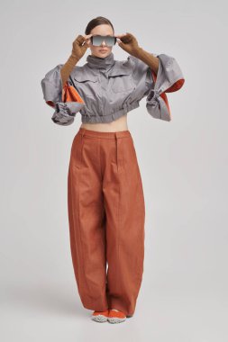 Gri ceketli, geniş kahverengi pantolonlu ve gri arka planda aynalı güneş gözlüklü güzel bir kız modelin tam boy portresi. Fütürist moda tarzı. Yüksek moda.