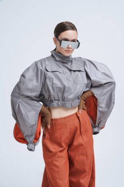 Gri ceketli, geniş kahverengi pantolonlu ve gri arka planda aynalı güneş gözlüklü güzel bir mankenin portresi. Fütürist moda tarzı. Yüksek moda.