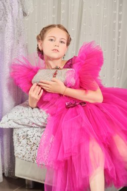 Lüks bir fuşya elbisesi içinde poz veren ve gece elbiselerinden oluşan bir salonda gümüş bir el çantasıyla küçük tatlı bir bayanın portresi. Akşam saç stili. Çocuklar için Yüksek Moda.