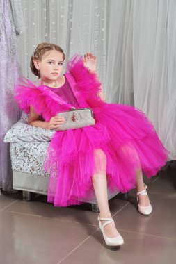 Lüks bir fuşya elbisesi içinde poz veren ve gece elbiselerinden oluşan bir salonda gümüş bir el çantasıyla küçük tatlı bir bayanın portresi. Akşam saç stili. Çocuklar için Yüksek Moda.