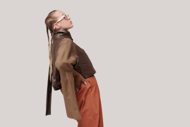 Eğilimler. Özel tasarım koleksiyonu. Tasarımcı kahverengi deri ceket, bluz ve turuncu geniş bacak pantolonuyla poz veren güzel bir manken kız. Stüdyo görüntüsü gri arka planda. Güneş gözlüğü. Metin için yer.