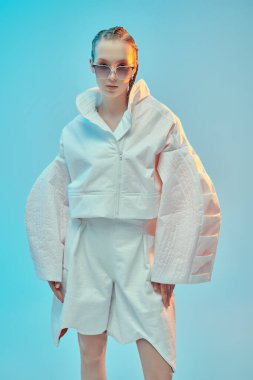 Beyaz ceketli, beyaz pantolonlu ve mavi arka planda büyük güneş gözlüklü güzel bir kadın model poz veriyor. Gelecekçi tarzı. Yüksek moda.