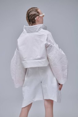 Yüksek moda. Gri stüdyo arka planında beyaz tasarımcı kıyafetleri ve zarif güneş gözlükleriyle güzel bir kadın model poz veriyor. Örgülü saç stili.