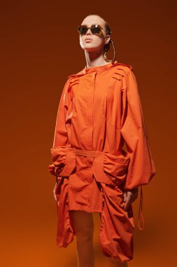 Portakal rengi pelerinli ve son moda güneş gözlüklü şık bir manken kız turuncu stüdyo arka planında poz veriyor. Yaz koleksiyonu. Saç stili örgülü. Yüksek Moda Çekimi.