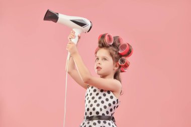 Zarif puantiyeli elbisesi ve kafasında bigudileri olan güzel bir kız partiye gidip saç kurutma makinesi kullanıyor. Pembe stüdyo geçmişi. Pin-up stili. Çocuk modası. Metin için boşluk.