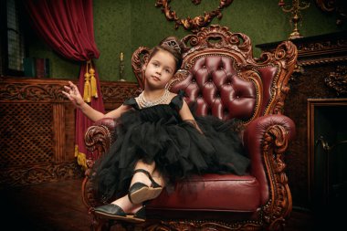 Kabarık siyah bir elbise giymiş, akşam saç stili ve takıları klasik bir koltuk ve gülümsemeyle süslenmiş güzel bir kızın tam boy portresi. Lüks daireler. Kızlar için akşam modası.