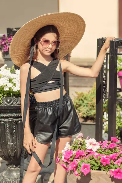 Zarif, geniş kenarlı bir şapka ve güneşli bir yaz gününde açık havada moda olan güneş gözlüklü güzel bir kızın portresi. Çocuk modası. Şehir sokaklarında küçük bir moda düşkünü.  