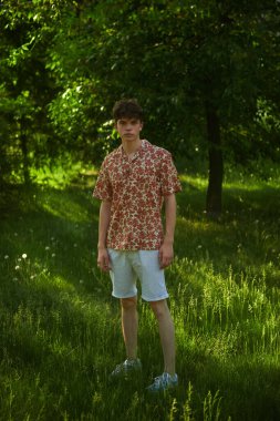 Yaz tişörtlü ve şortlu yakışıklı bir gencin uzun bir portresi yaz parkındaki ağaçların arka planına karşı. İnsanlar, hayat anları, duygular. Yaz zamanı..