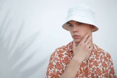 İnsanlar, hayat anları, duygular. Beyaz bir duvara karşı beyaz panama ve yazlık gömlekli modern bir gencin portresi. Sokak fotoğrafı. Yaz modası. Metin için yer.