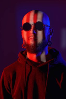 Yaşam tarzı. Dudağına piercing takan, siyah güneş gözlüğü takan ve siyah kapüşonlu vahşi, sakallı bir adamın fotoğrafı. Stüdyo görüntüsü koyu mor arkaplanda karışık kırmızı ve mavi ışıkta.