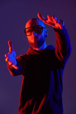 Kel, sakallı, dudak piercingli, siyah tişörtlü ve büyük gözlüklü vahşi bir adamın karışık renkli görüntüsü. Stüdyo mor arkaplan.