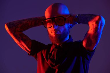 Kel, sakallı, dudak piercingli, siyah tişörtlü ve büyük gözlüklü vahşi bir adamın karışık renkli görüntüsü. Stüdyo mor arkaplan.