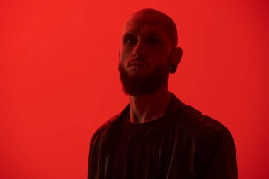 Dudaklarında ve kulaklarında piercingler olan sakallı, vahşi bir adam kırmızı stüdyo arka planında siyah deri bir ceketle poz veriyor. Metin için boşluk.