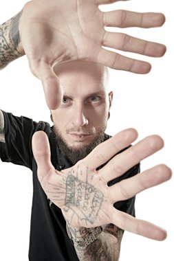 Siyah bir papaz gömleği giymiş, vahşi, tıraşlı bir adam ellerini bir dövmeyle gösteriyor. Antisosyal davranış, protesto ifadesi. Beyaz stüdyo geçmişi.