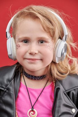 Küçük DJ kız. Parlak makyajlı, siyah deri ceketli ve kulaklıklı, kırmızı stüdyo arka planındaki pop ve rock müzisyeni gibi şirin bir kızın portresi. Çocuk modası.