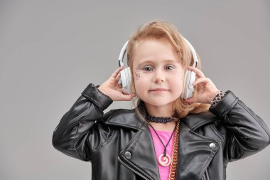Parlak makyajlı, modaya uygun kıyafetleri ve aksesuarları olan sevimli bir kız ve rock müzisyeni kulaklıkla müzik dinliyor. Gri stüdyo geçmişi. Çocuk modası. DJ çocuk.