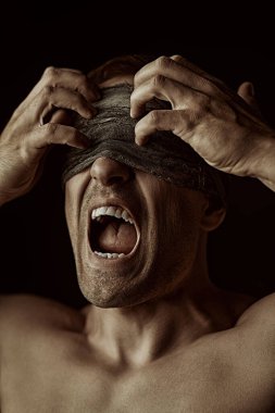 Çıplak gövdeli, iri yarı, güçlü bir adam sargı beziyle gözlerini kapatır ve bağırır. Siyah arka planda stüdyo portresi. Psikolojik resim.