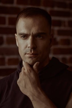 Siyah kapüşonlu güçlü bir adamın yakın plan portresi, odaklanmış ve yoğun bir şekilde kameraya bakıyor. Arka planda tuğla duvar var. Halk konsepti. Erkekler, erkeklik ve güç.. 