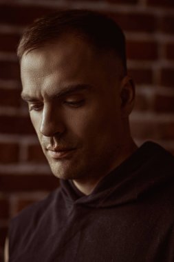Siyah kapüşonlu güçlü bir adamın yakın plan portresi. Odaklanmış ve sakince aşağıya bakıyor. Arka planda tuğla duvar var. Halk konsepti. Erkekler, erkeklik ve güç..