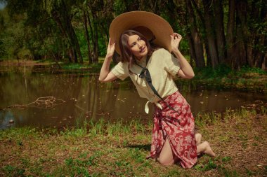 Mutlu ve huzurlu yaz havası. Güzel, romantik bir Asyalı kız, resimli bir yaz parkındaki göletin kıyısında oturuyor. Yaz modası.