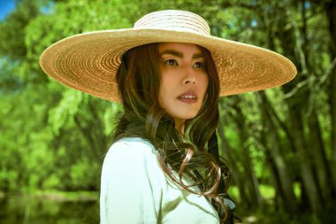 Geniş kenarlı hasır şapkalı, romantik bir Asyalı kız bir yaz parkında hayali bir göletin yanında duruyor.. 