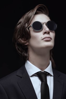 Erkek modası ve tarzı. Şık saç stili olan, zarif siyah klasik bir takım elbise, beyaz gömlek ve zarif güneş gözlüğü takan bakımlı bir gencin portresi. Siyah arkaplan. 