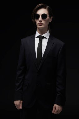 Şık saç stili olan, zarif siyah klasik bir takım elbise, beyaz gömlek ve zarif güneş gözlüğü takan bakımlı bir gencin portresi. Siyah arka plan. Erkek modası ve tarzı.