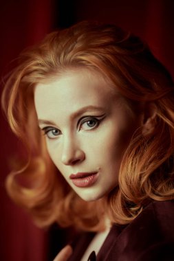 Kadın güzelliği ve tarzı. Pahalı kadife ceketli, loş ışıklı bir odada kızıl saçlı çekici bir genç kadın. Moda ve kadınlık. Oklarla ve saç stiliyle zarif bir makyaj..