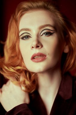 Kadın güzelliği ve tarzı. Pahalı kadife ceketli, loş ışıklı bir odada kızıl saçlı çekici bir genç kadın. Moda ve kadınlık. Oklarla ve saç stiliyle zarif bir makyaj..