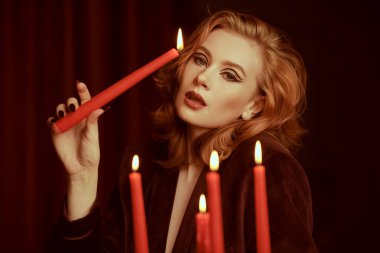 Kahverengi kadife ceketli, koyu renk bir odada mumları olan kızıl saçlı hoş bir bayan. Zarif saç stili ve oklu makyaj. Kadın Güzelliği ve Moda.