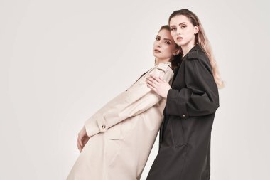 Beyaz bir stüdyo arka planında moda trençkotlarla poz veren iki şık güzel kız mankeni. Kadın modası ve güzelliği.