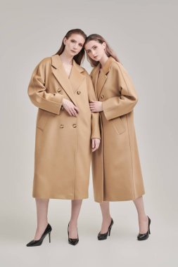 Yarı sezon kadın modası. Uzun bej keçeli ceketlerle poz veren iki güzel manken kızın tam boy portresi. Beyaz stüdyo geçmişi.